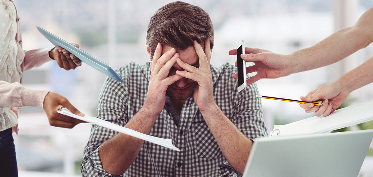 Man stressed with head down and hands on forehead while co-workers hand him papers, phone, pencil, and notebook at once.